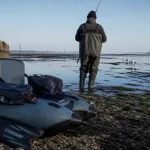 Float tube mer gris-bleu au bord de l’eau, pêcheur en waders et palmes.